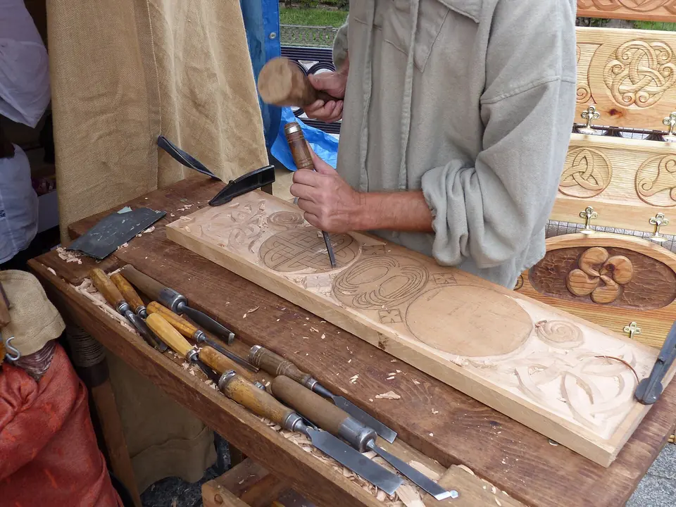 Mains d'un artisan sculptant un panneau de bois au ciseau, avec des gouges alignées sur l'établi.