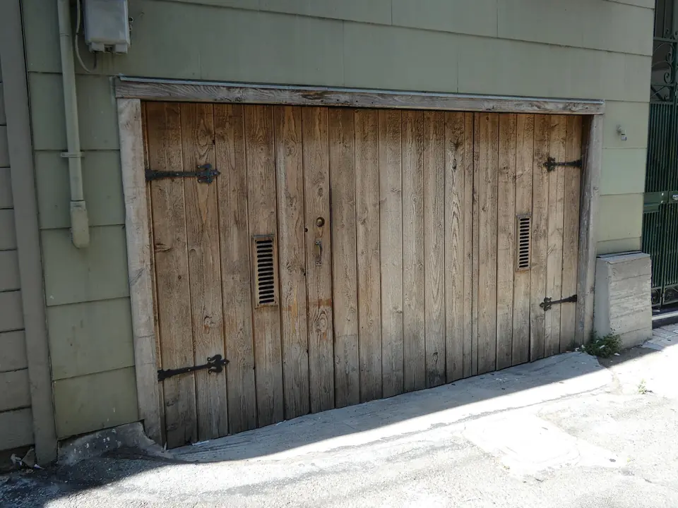 Porte de garage en planches de bois brut patiné, avec pentures métalliques noires, encastrée dans une façade à bardage vert.